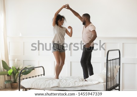 Young romantic african american couple wearing pajamas dancing on bed at home, happy active mixed race husband and wife enjoying funny party on honeymoon together in the morning in bedroom interior