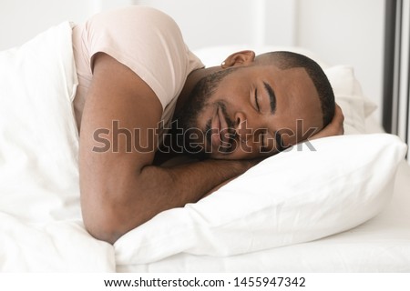Serene calm handsome young black man sleeping well alone on orthopedic soft pillow under warm duvet, african american guy lying asleep in comfortable cozy bed enjoy good night peaceful healthy sleep Royalty-Free Stock Photo #1455947342