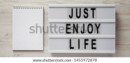 A lightbox with 'Just enjoy life' words, blank notepad on a white wooden surface, top view. Copy space.