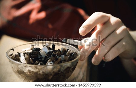 The man is holding a cigarette and flicking the ashes into a dirty glass ashtray full of cigarette butts. Ashtray - the personification of the abomination of Smoking.