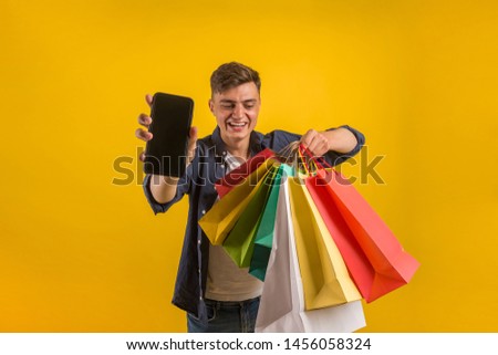 Handsome guy with shopping bags is using a mobile phone and smiling. Portrait of a smiling man holding shopping bag over yellow background and looking at camera