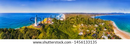 Top of headland with Byron Bay lighthouse high above Pacific ocean coast on a sunny day in elevated aerial panorama facing inland over sandy beaches. Royalty-Free Stock Photo #1456133135