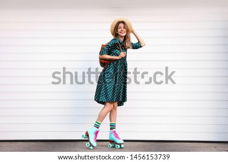 Happy stylish young woman with vintage roller skates, hat and backpack near white garage door on street