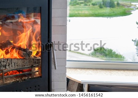 Cozy home with black safe fireplace with burning orange flames from wood - it’s near the window with beautiful picturesque view on the lake, meadow, forest