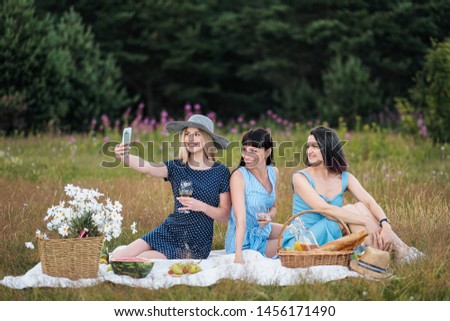 Three young women, in blue dresses and hats sit on plaid and take pictures on smartphone. Outdoor picnic on grass. Delicious food in picnic basket and wine. Watermelon, grapes and bouquet of daisies.