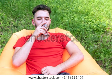 Caucasian traveler drinks wine at a picnic resting in the forest.A guy with a beard in a red T-shirt is lying on an orange inflatable Lamzatz sofa.