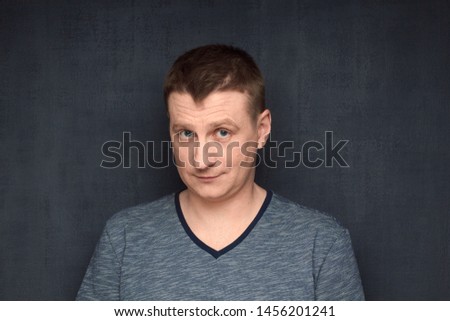 Studio close-up portrait of genial caucasian fair-haired man, wearing t-shirt, smiling kindly and and looking naively from under forehead at camera, making innocent face, against gray background Royalty-Free Stock Photo #1456201241