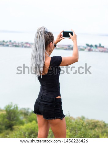 beautiful tanned caucasian female with blonde afro-braids in black clothes making photos with a smartphone from the cliff of the lake