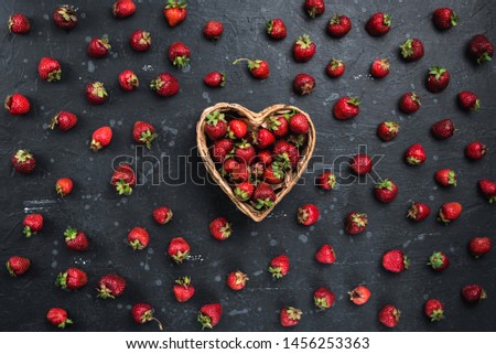 
A basket in the form of a heart with ripe strawberries among the juicy berries of ripe strawberries on a dark concrete surface. Dark background. Top view.