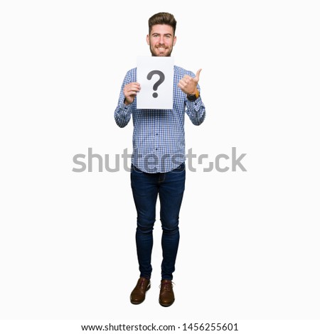 Young handsome man holding paper with question mark happy with big smile doing ok sign, thumb up with fingers, excellent sign