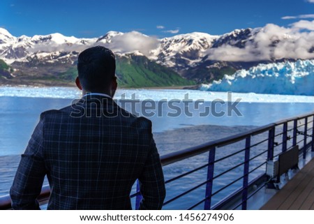 Businessman in suit is looking on Hubbard Glacier. Open deck on a cruise ship and amazing view on snow peaks of the mountains and white clouds. Juneau, Alaska. Blue ice on background.