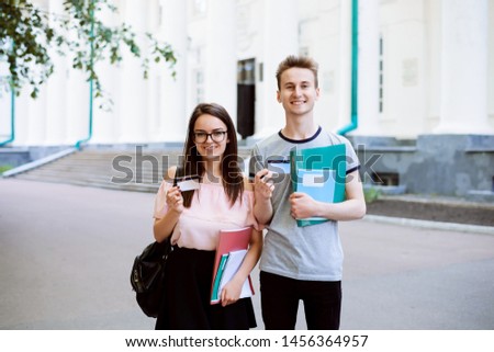 Two best friends smiling, holding credit cards and showing them to the camera in front of old university. Happy students use advantages of electronic cards in everyday life: buy goods, pay bills