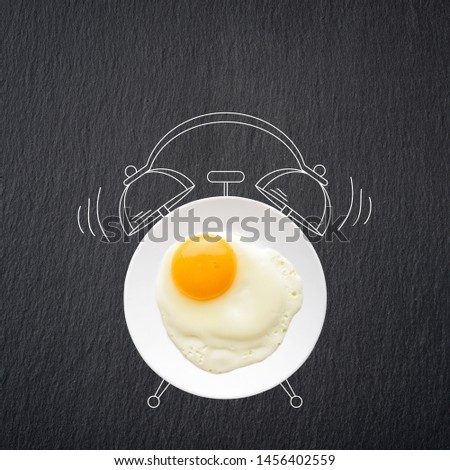 Plate with sunny side up fried egg and sketch of alarm clock on black slate background, top view