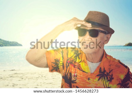 Picture of old man wearing summer clothes and hat while looking far away at sea and standing on the beach