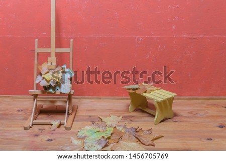 Traditional easel stand with dried leaves, small wooden bench, and dried leaves on wooden floor and red painted wall in studio. Autumn decoration.