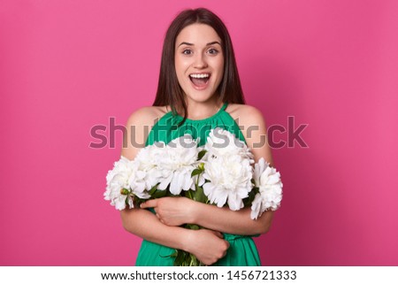 Indoor studio shot of emotional charismatic female standing isolated over pink background, looking directly at camera, holding big bouquet of white peonies, opening her mouth and eyes widely.