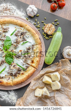 Pizza with zucchini, meat, cream cheese on the table in the restaurant. Garlic, zucchini, champignons and pieces of cheese near on the table. Vertical image. Natural light. Black background. 