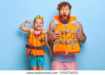 Father and daughter in safety lifejackets going to have extreme activity on water. Shocked bearded ginger man stares at camera, cheerful little girl shows biceps, have summer rest together. Family Royalty-Free Stock Photo #1456756055