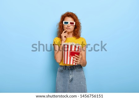Indifferent red haired young lady wears three dimensional glasses, watches attentively documentary film, holds big bucket of popcorn, has serious look, poses indoor. People and leisure concept