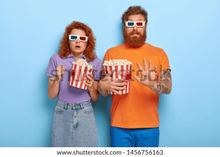 Image of scared red haired boyfriend and girlfriend eat popcorn and watch horror movie, gesture actively, have bated breath from fear, wear 3d glasses in cinema. Frightened couple enjoy excting film Royalty-Free Stock Photo #1456756163