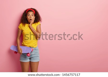 Studio shot of surprised dark skinned model with curly hairstyle, has frightened look, carries purple longboard, likes skateboarding, reacts on sudden news. Modern youth hobby and recreation