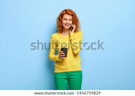 Portrait of pleasant looking foxy woman drinks coffee, calls via smartphone, makes order, books table restaurant, has fun during hilarious chat, isolated over blue background. People and communication