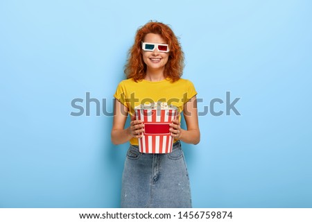 Photo of happy red haired millennial female wears 3d glases, eats tasty popcorn, enjoys watching favourite film, smiles gently, dressed in fashionable outfit. Pretty viewer with junk food in cinema