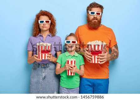Bored ginger father and mother come on cinema together with small daughter for watching new cartoon in 3d, wears dimension stereo glasses, eat popcorn. Glad child spends time with loving parents