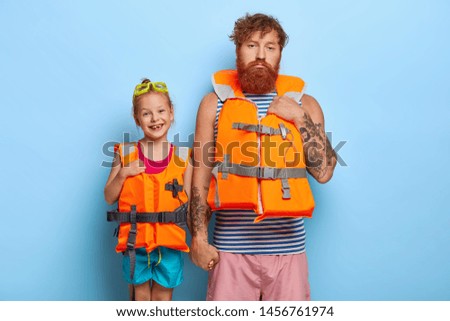 Image of lovely glad little girl wears swimming goggles and orange lifejacket, holds hand of dad. Sad tired lifesaver gives training lesson for small kid. Ginger family spend summer rest actively