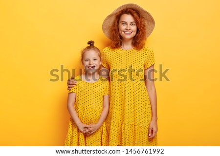 Studio shot of ginger female in stylish straw hat and dress embraces small sister, buy same clothes, smile happily, look alike, have good relationship, isolated on yellow background. People, happiness Royalty-Free Stock Photo #1456761992