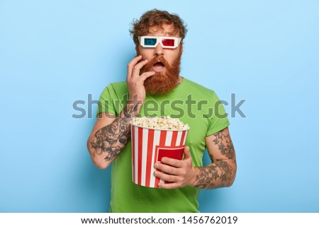Emotional stupefied red haired man stares at camera through 3d cinema glasses, wears casual green t shirt, holds bucket of fresh popcorn, isolated on blue background. Shocked spectator indoor