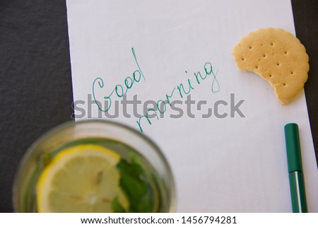 The inscription on a white sheet of paper - good morning. Nearby is a pen. Next to a glass of green tea, bitten by cookies.