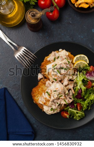 Fried cod, served on potato pancakes with mushrooms sauce and salads. Portion on a black plate. Dark background.