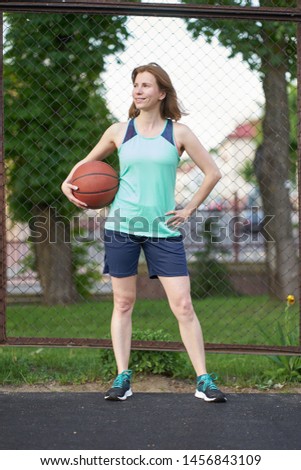 Redhead girl standing on the outdoor basketball court, holding ball, and smiling