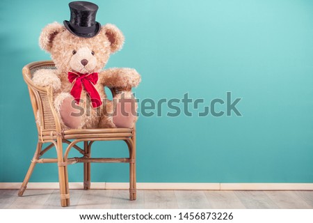 Retro big Teddy Bear toy in magician black cylinder hat sitting on old aged wooden chair front mint blue wall background. Vintage style filtered photo