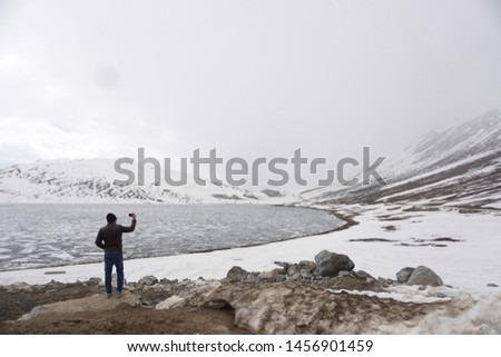 A person/tourist seen from the back taking photographs on his cellphone at a frozen snowy lake in Northern Pakistan at Sheohosar Lake, Deosai National Park.  Perfect for a travel concept.