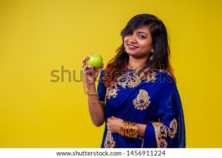 Close up portrait of a young Indian brunette woman with traditional blue sari in studio in yellow copy space background biting an apple in her hand.