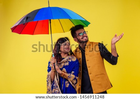 beautiful indian woman in traditional blue india sari and romantic man in kurta together standing under multicolor rainbow umbrella in yellow studio background .