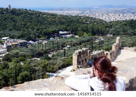 A girl with brown hair takes pictures of the Odeon. Ancient Greek ruins, ruins amidst lush green grass. Acropolis, Athens, Greece on a hot sunny day.