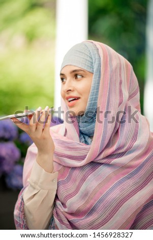 Young female in hijab recording voice message on smartphone outdoors