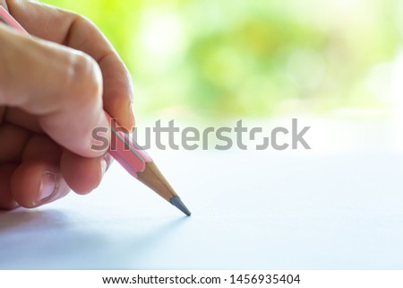 Woman's left Hand holding multicoloured pencil, writing letter on white paper, Bokeh green garden background, Notebook, Close up & Macro shot, Selective focus, Communication, Stationery concept