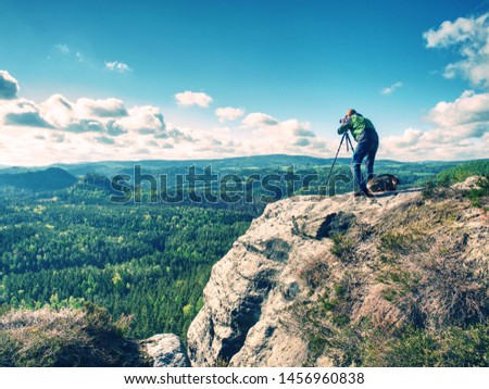 Adult caucasian photographer with telephoto Lens taking Pictures  of midday hilly landscape. Artist is using camera on Tripod.