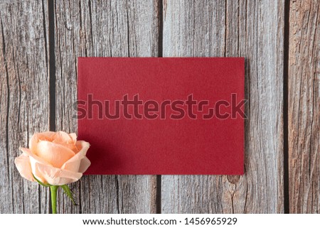 A piece of bright red paper with a single peach color rose on brown wooden background.   Mock up invitation card. Top view. Flat design.
