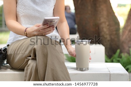 Hand of a woman grabbing reusable coffee cup, tumbler while texting message in her smart phone. Environmental friendly, eco living, No one-time use plastic, No straw, Urban leisure, Zero waste concept Royalty-Free Stock Photo #1456977728