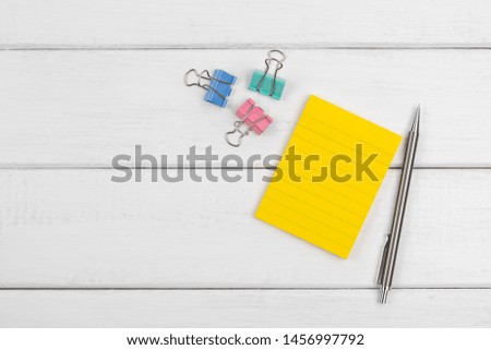 Top view of a yellow blank sticky note with pencil and paper clips on white wooden background