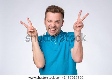 young smiling caucasian man in blue polo shirt showing the victory sign and looking at the camera. Studio shot
