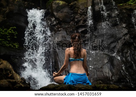 Young Caucasian woman meditating, practicing yoga and pranayama with gyan mudra at waterfall in Bali. Yoga retreat in Bali. Healthy life. View from back. Pengibul waterfall. Ubud, Bali, Indonesia
