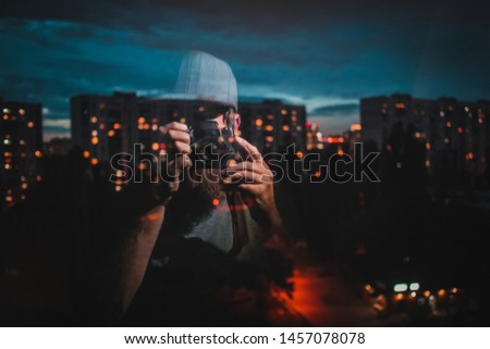 Reflection of a hipster photographer with a baseball cap on a picturesque evening cityscape of Kiev city blocks with lights on in the apartments. Lights in soft focus around photographer
