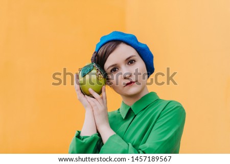 The concept of fashionable summer, blue and green colors. Stylish woman in a blue beret and a green shirt is standing against the orange wall with various emotions. portrait with the apple close up