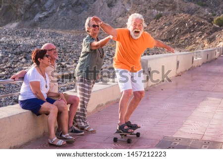group of seniors and mature people at the beach have fun looking at old man riding a skateboard and laughing with scare face - woman touching the man
